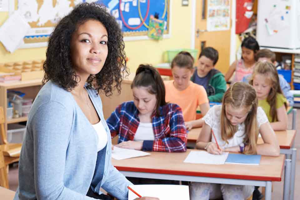 A female tutor is smiling by looking at the camera and at background it appears to be students are engage in their botany homework and assignment