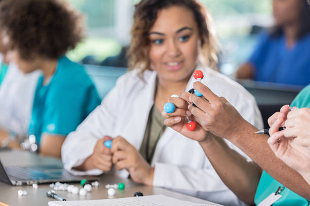 A group of students in lab coats and scrubs are sitting at a table with laptops and molecular models, working on their molecular biology homework. One student, in a white lab coat, looks on as another holds a colorful balls-and-sticks molecular model for their molecular biology assignment help session.