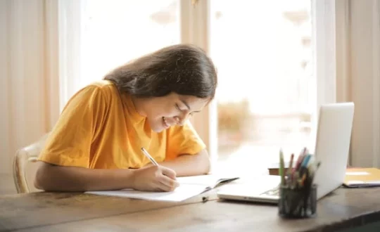 A young woman with long hair is sitting at a wooden table, smiling and writing in a notebook. She is wearing a yellow shirt and has an open laptop in front of her, along with a pen holder filled with pens and pencils. Sunlight streams through the window as she seeks bioinformatics assignment help online.