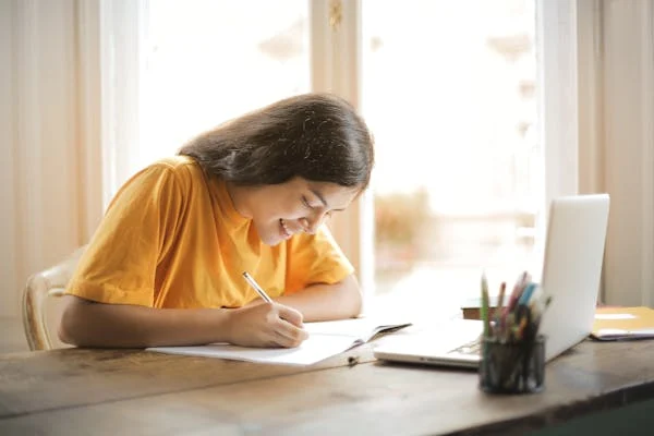 A young woman with long hair is sitting at a wooden table, smiling and writing in a notebook. She is wearing a yellow shirt and has an open laptop in front of her, along with a pen holder filled with pens and pencils. Sunlight streams through the window as she seeks bioinformatics assignment help online.