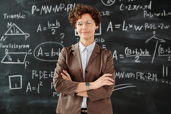 A woman with short curly hair wearing glasses and a brown blazer stands confidently with arms crossed in front of a chalkboard filled with advanced math equations and geometric figures