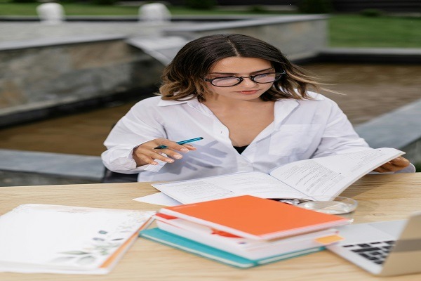 A student with glasses is seated at an outdoor table, focused on her algebra homework. She holds a pen in her right hand and a book in her left. The table is covered with several books and papers, including an assignment, with a laptop nearby. A stone fountain is visible in the background.