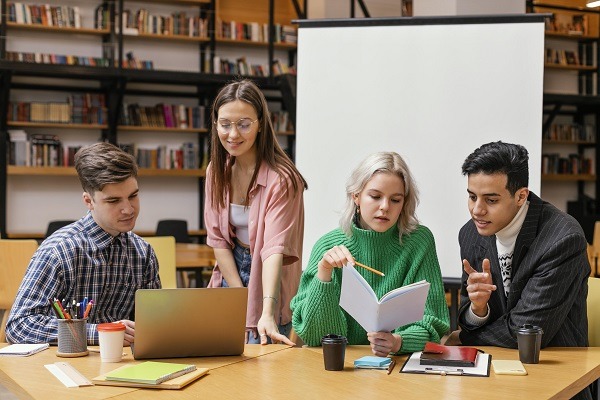 A group of four students sitting in a library with laptop and notebooks appears to be engage in analytical homework and assignment