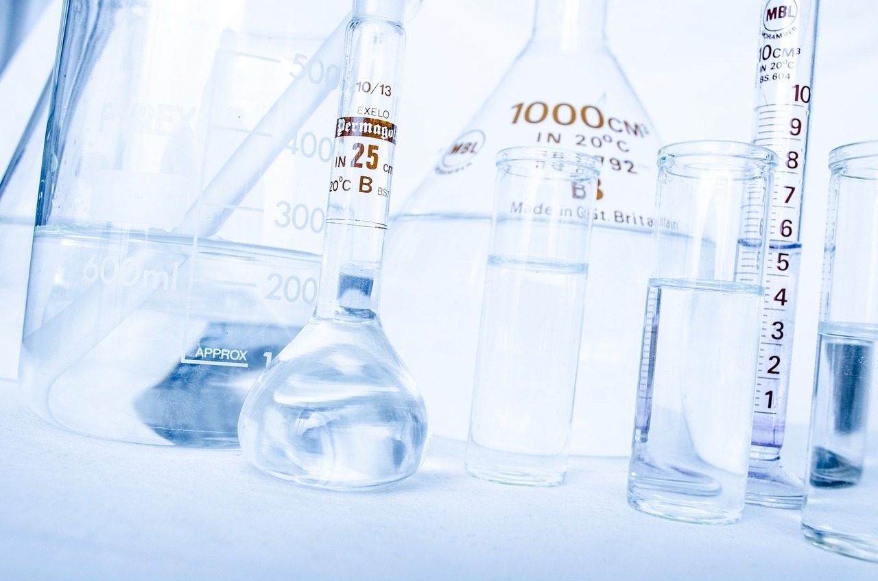 A close-up photo of various laboratory glassware used in analytical chemistry, including a 1000 mL Erlenmeyer flask, a graduated cylinder, and multiple test tubes filled with clear liquid, arranged on a white surface. The equipment is marked with precise measurement labels in milliliters.