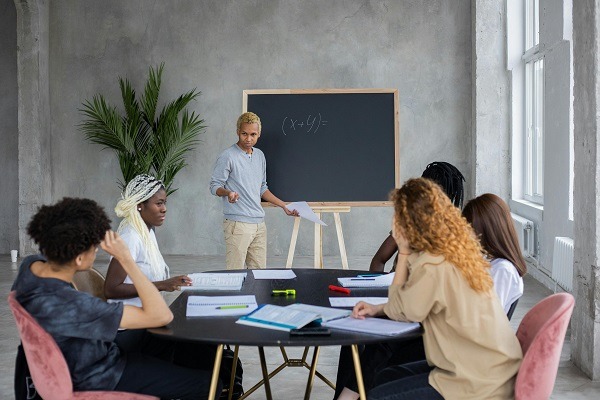 A person stands and points to a chalkboard with a mathematical equation while five others, seated around a circular table, look on. The table is scattered with economics homework, notebooks, and papers, and a large plant is in the background.