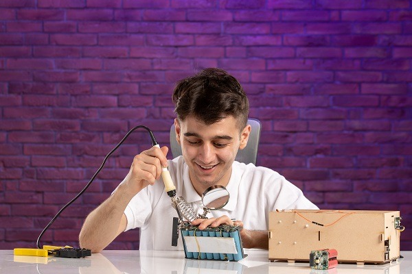 A person is smiling while soldering a circuit board for their electricity and magnetism project. They are seated at a white desk with a yellow multimeter and other electronics. The background features a purple brick wall