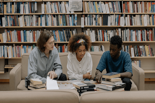 Three people are sitting on couches in a library, surrounded by piles of books. They appear to be engaged in a discussion or study session, possibly tackling finance homework and assignments while looking at a document. The background is filled with bookshelves