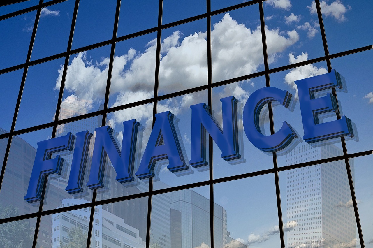 A reflective glass building facade prominently displays the word "FINANCE" in large, bold, blue letters. The sleek reflection captures drifting clouds and towering skyscrapers, underscoring its setting in a bustling urban financial district