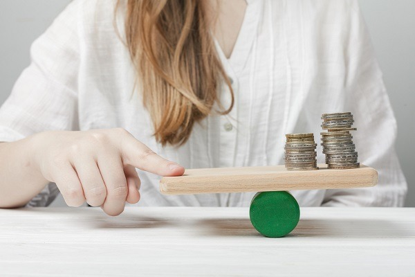 A female is balancing a wooden plank with their finger, demonstrating the principles of gravitation. The plank has a green cylindrical support in the middle and is topped with stacks of coins on one side. Wearing a white shirt, the individual carefully places their finger under the empty side of the plank. showcasing deep concept of gravitation assignment help