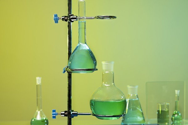 A laboratory setup with glass apparatus containing green liquid. A beaker and a flask are clamped vertically to a metal stand, illustrating principles of inorganic chemistry. Additional glassware with green liquid is visible in the background against a gradient yellow-green backdrop.