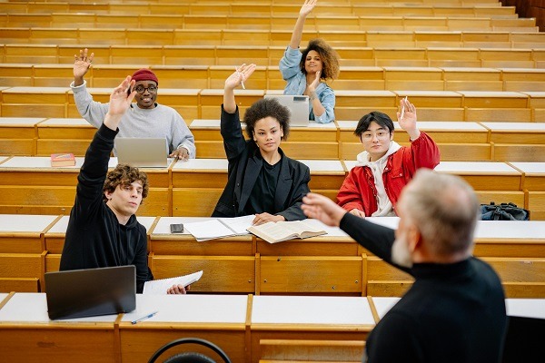 A group of students sitting in a lecture hall raises their hands to participate in an integral equation session. The professor, standing with his back to the camera, points towards them. Laptops and books are open on the desks.