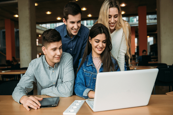 A group of four young adults, three men and one woman, are gathered around a laptop in a modern study environment. They are smiling and appear to be collaborating on a marketing project. One person is holding a tablet. The setting looks bright and open, perfect for an online marketing session