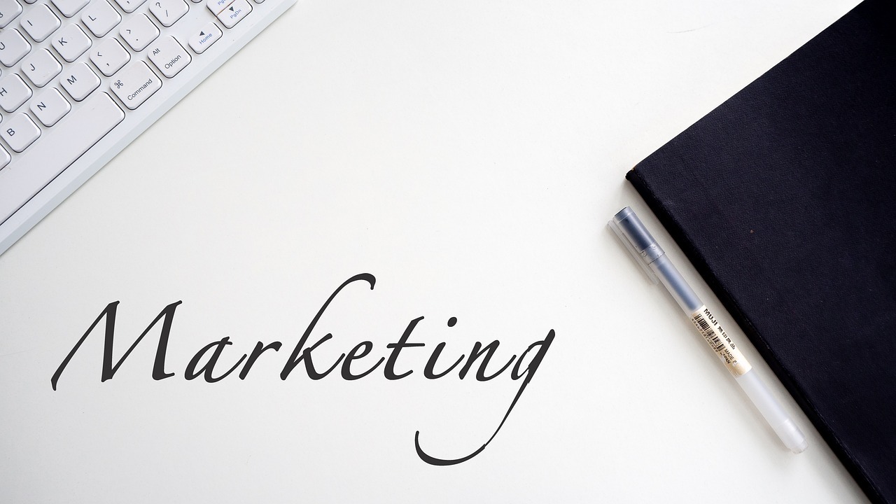 A white desk showcases the word "Marketing" elegantly written in cursive. To the left, a part of a keyboard is visible, while to the right, a black notebook with a clear pen resting on top completes the organized workspace