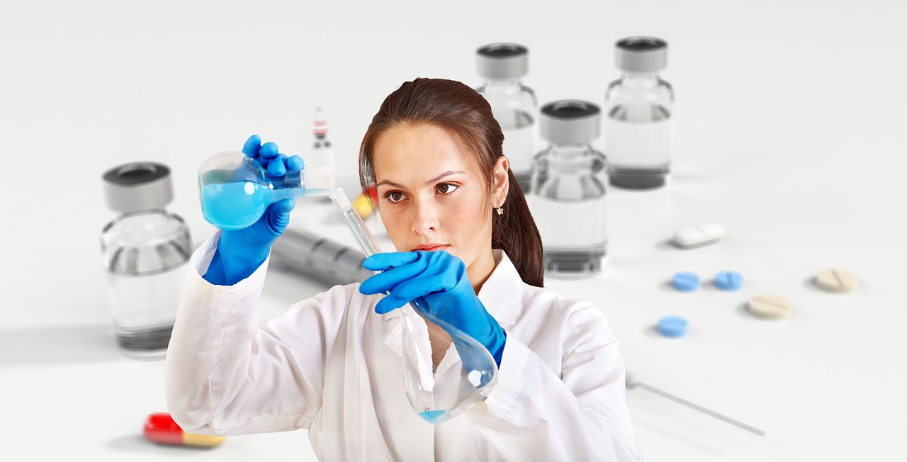 A scientist in a lab coat and blue gloves carefully pours a blue liquid from a flask into a test tube. Several vials, pills, and syringes are visible on the white surface around her, suggesting a medicinal chemistry laboratory setting.