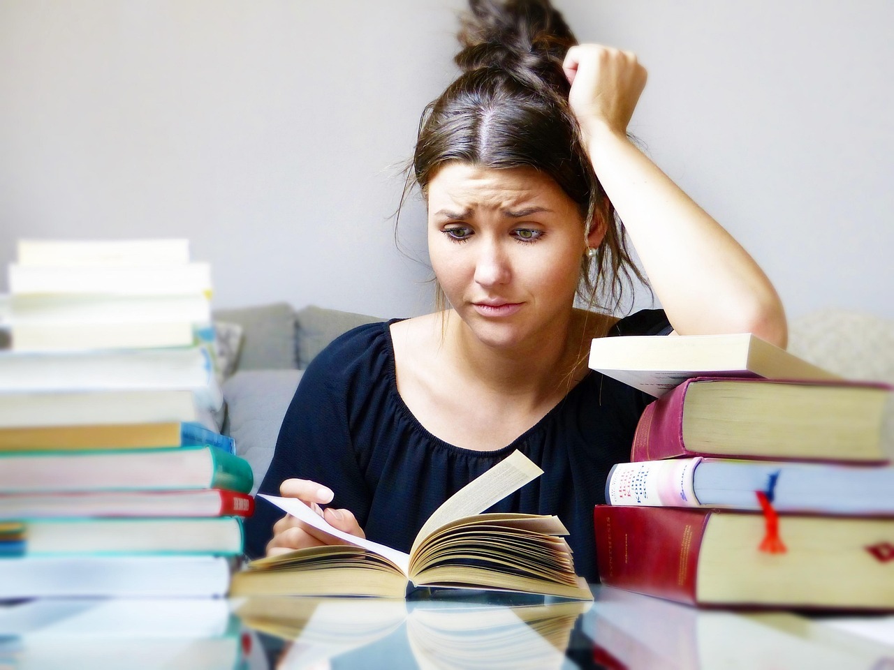 A young woman with a worried expression sits at a table, surrounded by multiple stacks of books. She holds an open book in her hand and rests her other hand on her head, appearing stressed or overwhelmed by the amount of nanotechnology homework and assignments