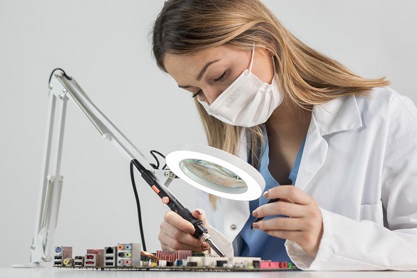 A student of nanotechnology wearing a lab coat and a face mask is using a soldering iron to work on an electronic circuit board under a magnifying lamp. She appears focused, potentially advancing the field of nanotechnology, while seated at a workbench filled with electronic components