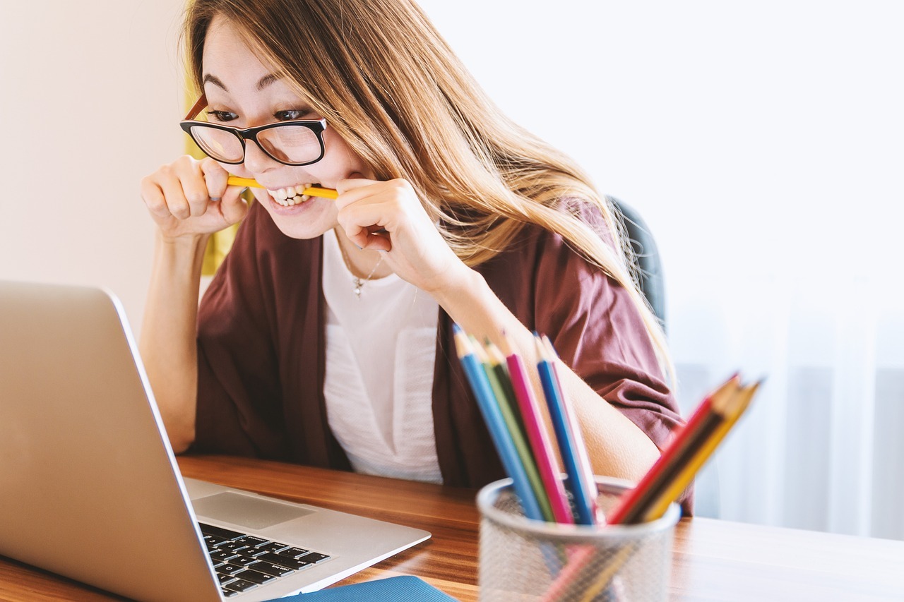 A female student with long hair, glasses, and a maroon cardigan is sitting at a desk, looking at a laptop with a frustrated expression. She is biting a pencil, likely struggling with her number theory homework. A container holding colored pencils and an assignment notebook are on the desk beside her.