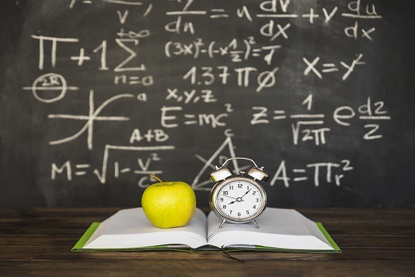 An open book with a green apple and an alarm clock on top sits on a table in front of a blackboard. The blackboard is filled with various number theory homework help equations and symbols written in chalk.