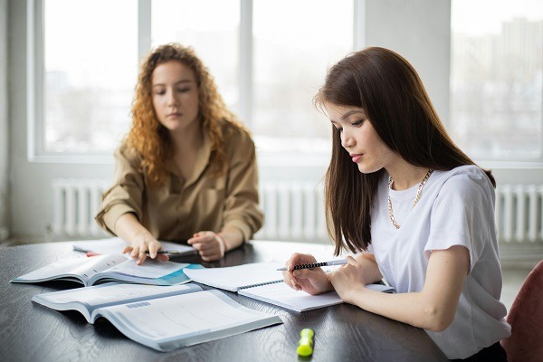 Two students are sitting at a table with books and notebooks. One, with brown hair, is writing in a notebook, likely working on her operations research homework help, while the other, with curly hair, is engrossed in a textbook. Both are focused. The background shows large windows letting in natural light.