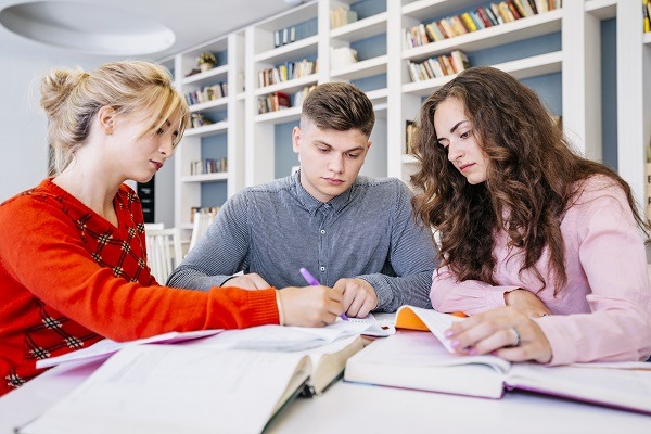 Three students sitting in a library writing on a notebooks with pens appear to be engaged in pathology homework and assignment.