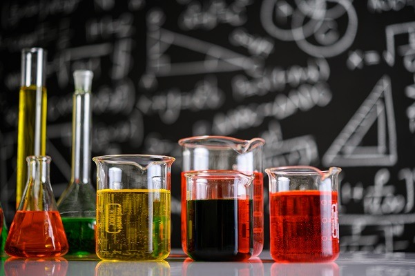 A collection of beakers and flasks filled with colorful liquids, including yellow, green, red, and dark red, are displayed on a reflective surface. The background features a blackboard adorned with various scientific formulas and geometric diagrams drawn in white chalk—an ideal setup for physical chemistry experiments