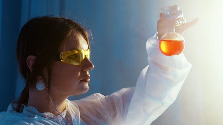 A female student wearing yellow safety goggles and a lab coat holds up a flask containing an orange liquid against a backdrop with blue lighting. They are inspecting the liquid closely, likely analyzing it for polymer chemistry research