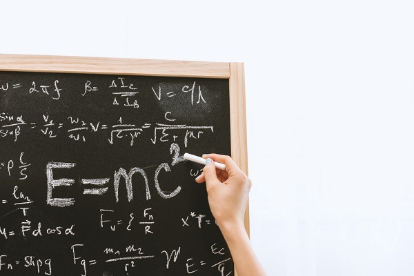 A hand holds a piece of chalk and writes on a blackboard filled with mathematical equations. The most prominent equation, "E=mc^2," is centered on the board, hinting at the complexities of quantum physics. The board is framed with a wooden border against a plain background