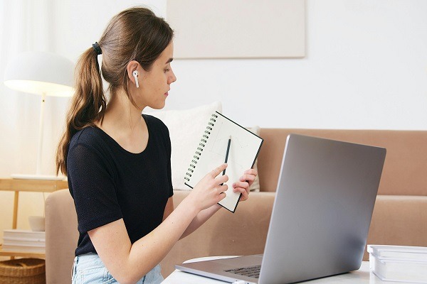 A student with brown hair tied back, wearing a black shirt and wireless earbuds, sits at a table with a laptop. She holds a spiral notebook and a pen, appearing to work on her semiconductor homework in a cozy, well-lit room.