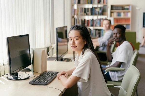 A young woman sits at a computer desk in a library, looking towards the camera. Next to her are computer monitors with thermodynamics homework displayed, and in the background, two other students are also working on assignments. Shelves filled with books line the walls of the well-lit room