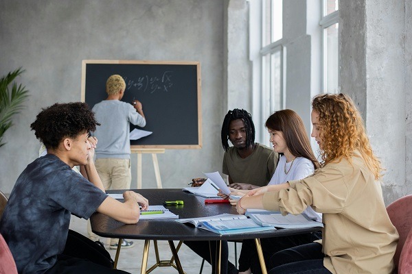 A group of five diverse people are in a thermodynamics session. Four of them are seated around a table with books and papers, discussing something with smiles and concentration. One person stands at a chalkboard, writing or drawing something in front of a window.