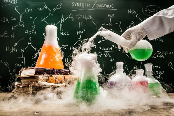 A scientist pours a green liquid from a test tube into a flask, causing smoke to rise. The table holds flasks with various colored liquids and old books, likely on organic chemistry. The chalkboard in the background is covered in chemical formulas and equations indicates organic chemistry assignment help concept