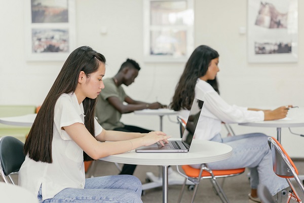 Four students are seated at desks in a classroom engaged in pharmacology assignment and homework. One student in the foreground is working on a laptop, while the others are writing with pens on paper. The classroom walls have framed pictures. The atmosphere appears studious and focused.