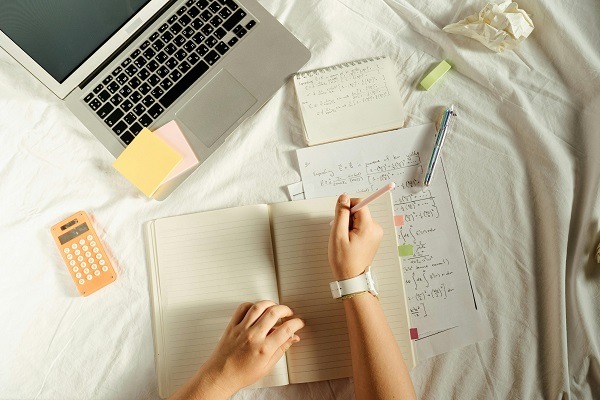 "A person is working at a cluttered desk that includes an open laptop, sticky notes, a notebook, papers with mathematical equations, a pen, and a calculator. The person's left hand holds a pen over a blank page in the notebook. Crumpled paper from working on probability homework is also seen nearby.