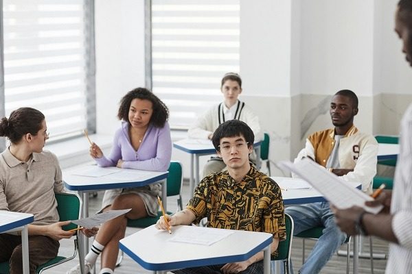 A diverse group of students sits at desks in a classroom setting, engaged and holding papers and pencils. Some look toward the instructor, who is partially visible on the right, while others are focused on their bioinformatics homework or looking around.