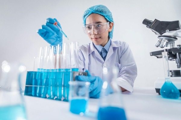 A biotechnician wearing a lab coat, safety glasses, gloves, and a hair cap is using a pipette to add liquid to one of many test tubes containing blue liquid. Laboratory equipment, including flasks and a microscope, is visible on the table.