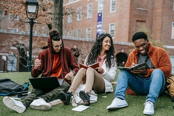 Three college students sit on grass in front of a brick building. They are chatting and smiling while looking at notebooks and a laptop. They are wearing casual clothing and appear to be studying a botany assignment.