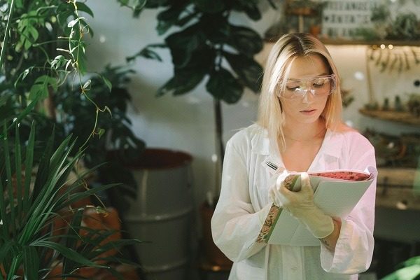 A botanical student wearing safety goggles and gloves reads from a notebook surrounded by green plants in a well-lit indoor space. She is dressed in a white lab coat and appears to be in a botanical or scientific environment.