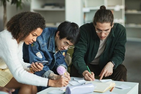 Three young adults are sitting closely together at a table, working on papers and notes. One is looking at a smartphone, another is writing, and the third is pointing at a paper with a pencil. They appear focused and engaged in their cellular biology assignment.