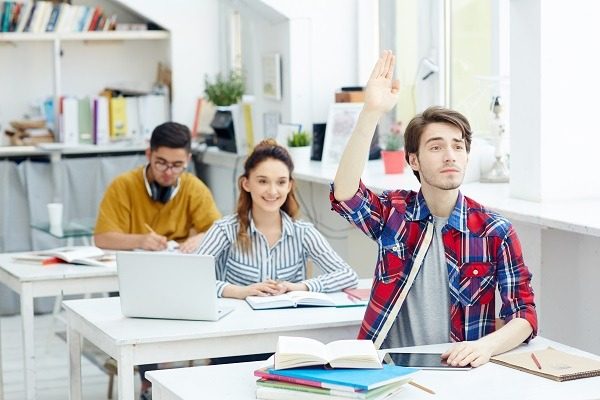 A classroom with three students seated at desks. The male student in the foreground wearing a red plaid shirt is raising his hand. A female student with long hair and a striped shirt is smiling, while another male student in a yellow shirt is focused on computational biology homework help on his laptop.