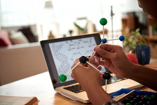 A person is holding a molecular model kit in front of an open laptop displaying a DNA sequence and chemical equations of computational biology homework help. The background shows a well-lit room with various objects, slightly out of focus.