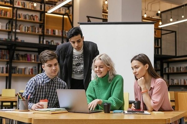 A group of four young adults gather around a laptop in a library. They seem engaged in epidemiology homework and assignment and are possibly collaborating on a project. Bookshelves and a large screen are visible in the background. One individual stands while the other three are seated.