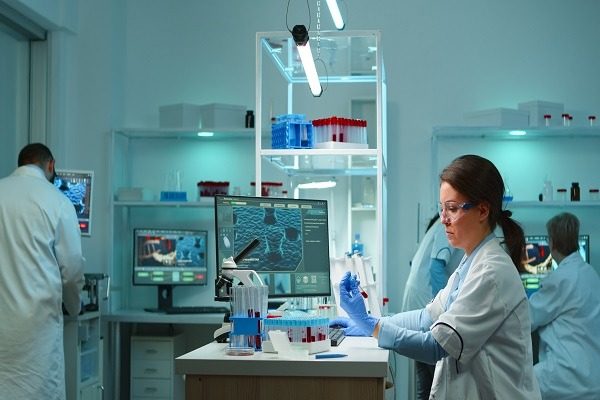 Epidemiology students in a lab conduct experiments with test tubes and a microscope. One student uses pipettes at a workstation with monitors displaying complex data. Shelves filled with lab equipment and samples are visible in the background. it depicts the concept of epidemiology assignment help