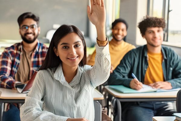 A young woman in a striped shirt raises her hand in a classroom, smiling at the camera. Three other students sit behind her at their desks, one wearing glasses and a plaid shirt, another in a yellow shirt, and the third in a mustard shirt. They all appear engaged in human anatomy physiology assignment help.