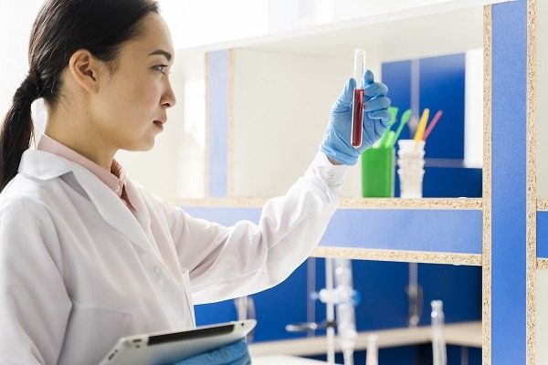 A microbiologist wearing a white lab coat and blue gloves examines a test tube containing a red liquid. They are holding a tablet in their other hand. The background shows laboratory equipment and shelves.