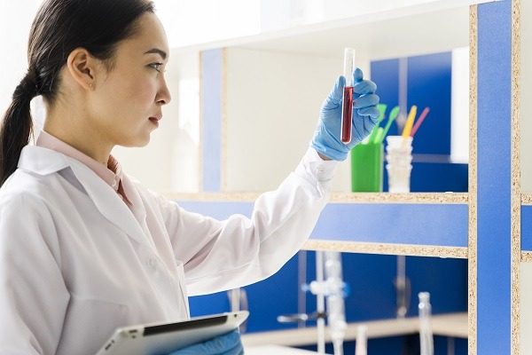 Microbiologist Analyzing Lab Sample A microbiologist wearing a white lab coat and blue gloves examines a test tube containing a red liquid. They are holding a tablet in their other hand. The background shows laboratory equipment and shelves.