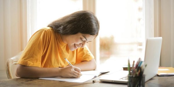 A girl with long hair wearing a yellow shirt is sitting at a desk, writing in a notebook pharmacy assignment with a pen. There is an open laptop and a cup holding colored pens on the desk. Sunlight is streaming through a large window behind them.