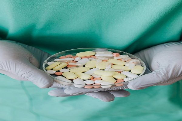 A pharmacy student wearing green medical scrubs and white gloves holds a transparent dish filled with assorted pills and tablets of various colors and shapes.