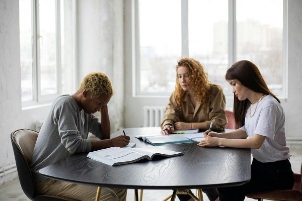 Three people are seated at a round table in a well-lit room, concentrating on their writing tasks. The person on the left appears stressed with their head resting on their hand, struggling with quantum physics homework, while the other two are focused on their assignments. A large book is open on the table