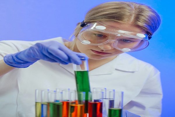 A pharmaceutics student wearing safety goggles and gloves is examining a test tube filled with green liquid. Several other test tubes containing various colored liquids are arranged in a rack in front of her. The background is lit with a blue hue. It depicts the concept of pharmaceutics assignment help