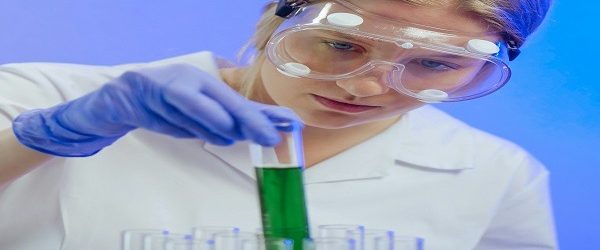 Pharmaceutics Student Analyzing Chemical Samples A pharmaceutics student wearing safety goggles and gloves is examining a test tube filled with green liquid. Several other test tubes containing various colored liquids are arranged in a rack in front of her. The background is lit with a blue hue. It depicts the concept of pharmaceutics assignment help